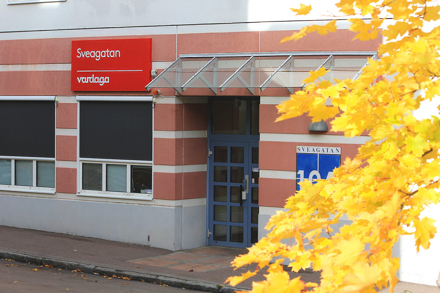 Vardagas huvudentré med en röd skylt ovan med texten "Sveagatan, Vardaga". Bredvid byggnanden syns gula löv från en lönn. 