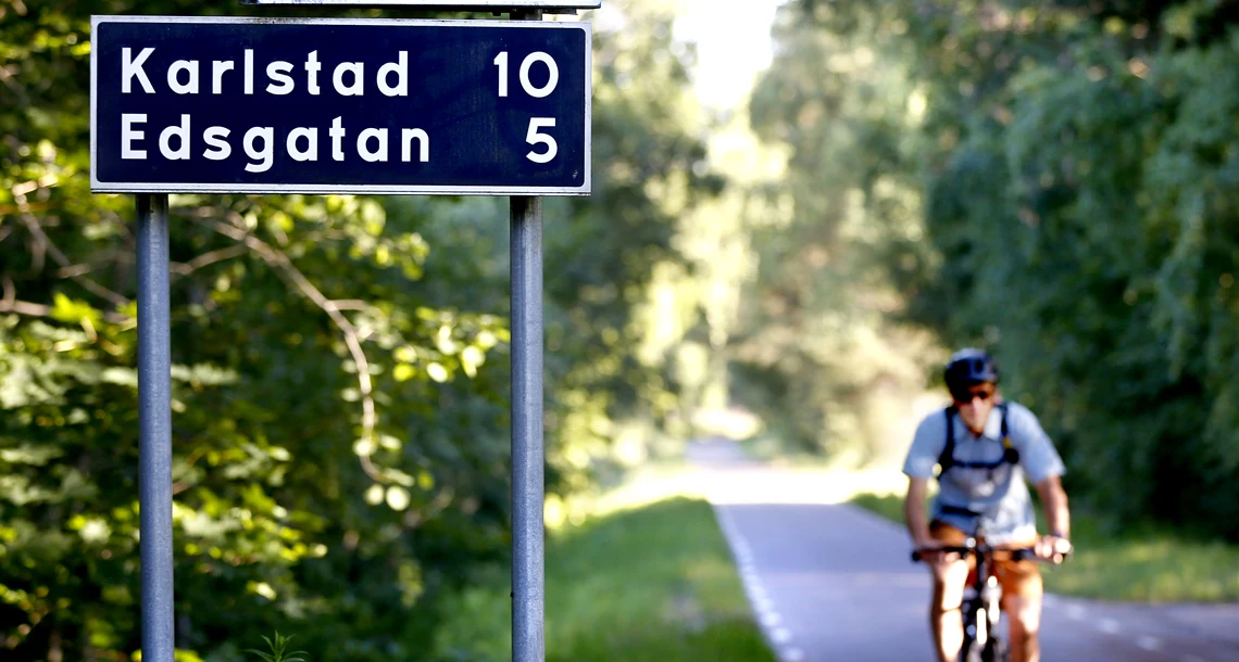 En cyklist som cyklar på en väg. I förgrunden finns skyltar där det står Ulvsby, Karlstad och Edsgatan samt antal kilometer till varje destination