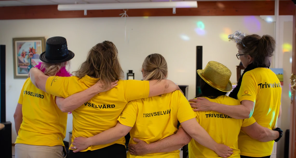 Fem trivselvärdar står med ryggen mot kameran och håller om varandra. På deras ryggar står det "trivselvärd".