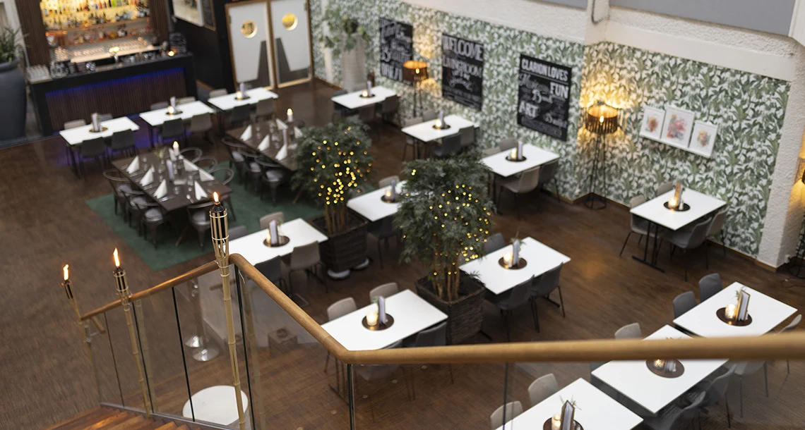 Ett ovanifrån perspektiv ner mot en större lokal där bord och stolar är utplacerade med luft emellan.