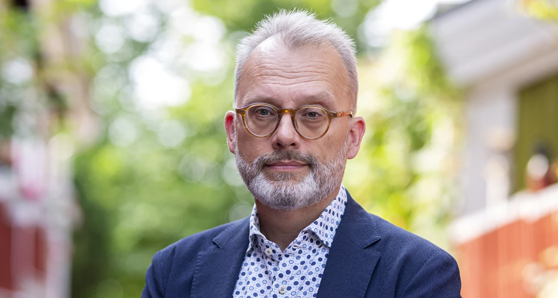 En porträttbild av en medelålders man med skägg. Han bär en skjorta och en blå kavaj.