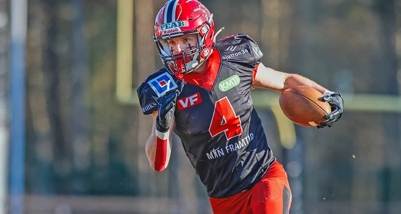 En kille i svarta och röda sportkläder och hjälm håller i en amerikansk oval fotboll