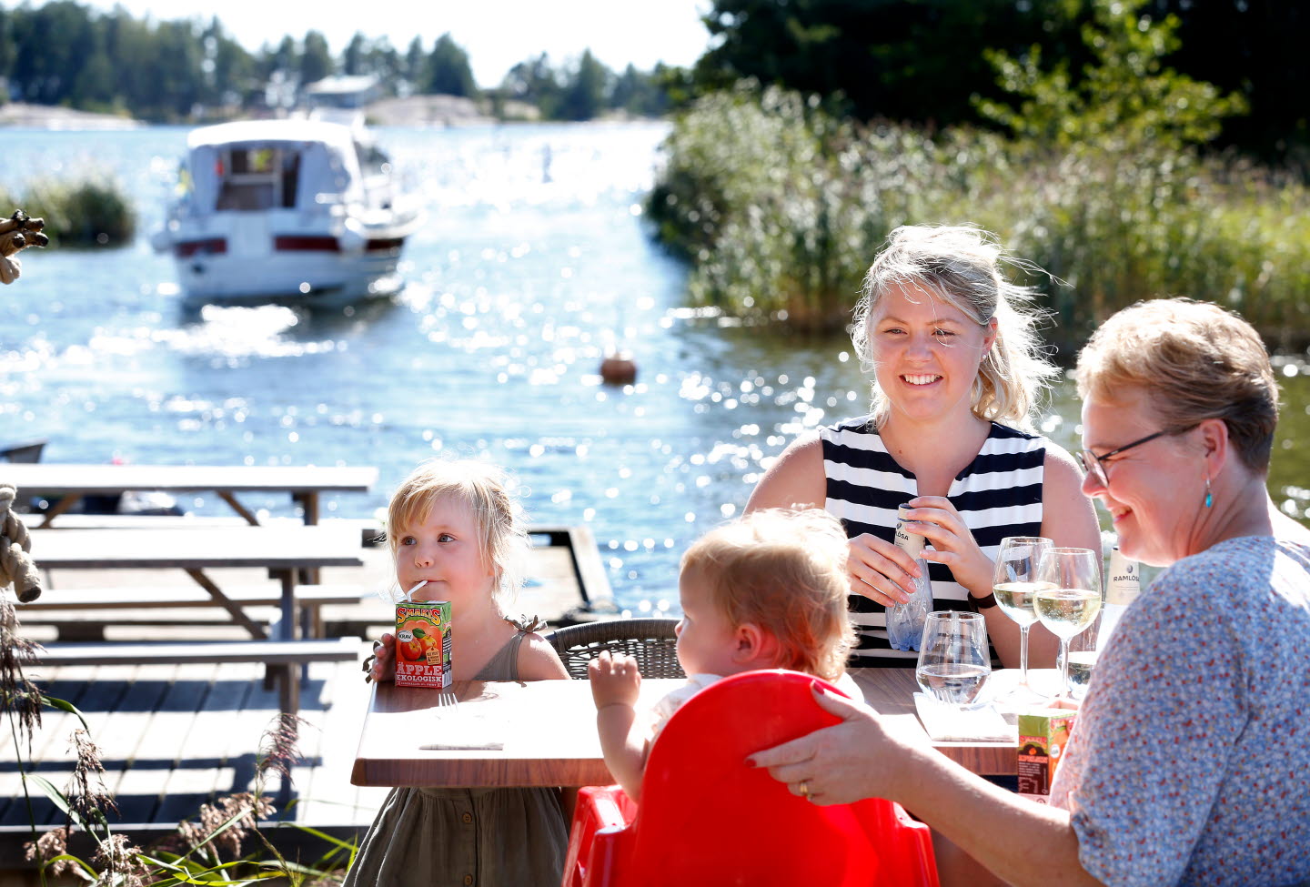 En kvinna, en äldre dam och två barn njuter av dryck vid ett bord. I bakgrunden ses en båt i vattnet