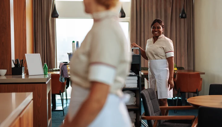 Två hotellanställda personer arbetar i ett konferensrum med en städvagn. 