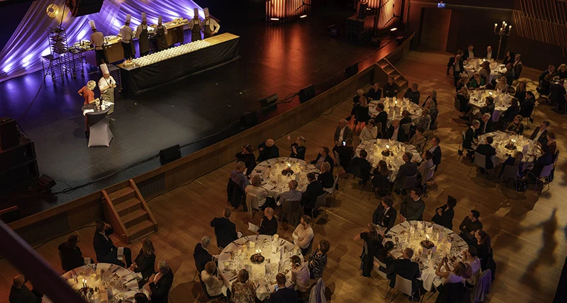 Flera runda uppdukade bord med matgäster framför en stor scen där två människor lagar mat.