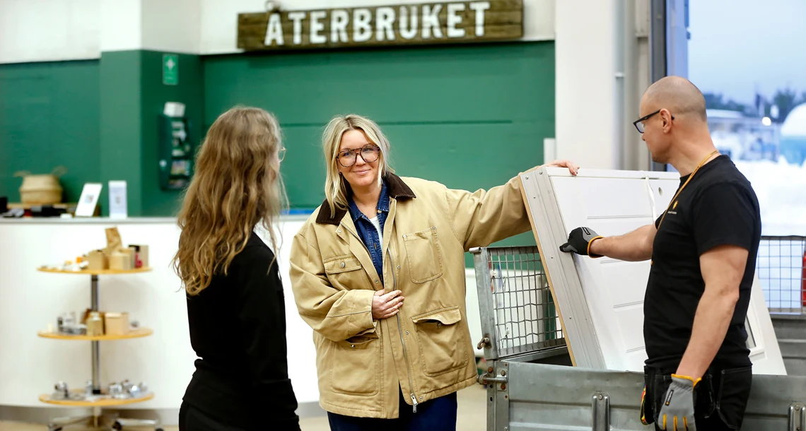 Tre personer i ett stort rum som liknar en lagerlokal. Det hänger en skylt med texten "återbruket" i bakgrunden.