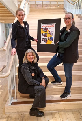 Tre kvinnor som står i en trappa och håller upp en affisch med bilder på  böcker. 