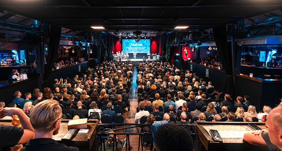En stor lokal med sittande människor på rader som tittar fram mot en scen med sittande människor på.