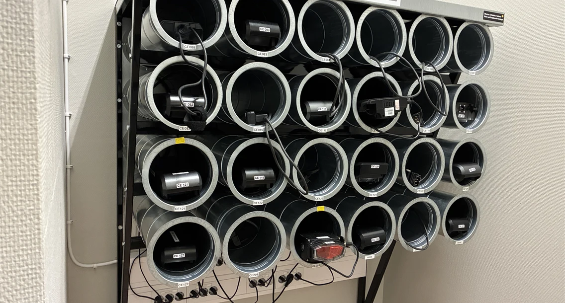 Laddställning för elcykelbatterier monterad på vägg i kommunalt laddrum. Batterier laddas i separata rör för ökad säkerhet och ordning.