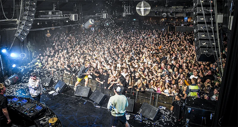 En konsert inomhus där tre människor står på scen med mikrofoner och glad människor i publiken sträcker upp sina armar i luften.