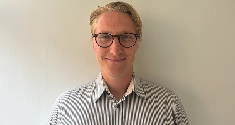 Porträttbild av en person framför en ljus vägg. Personen har ljust hår, glasögon och bär en ljus skjorta med smala, mörka ränder.