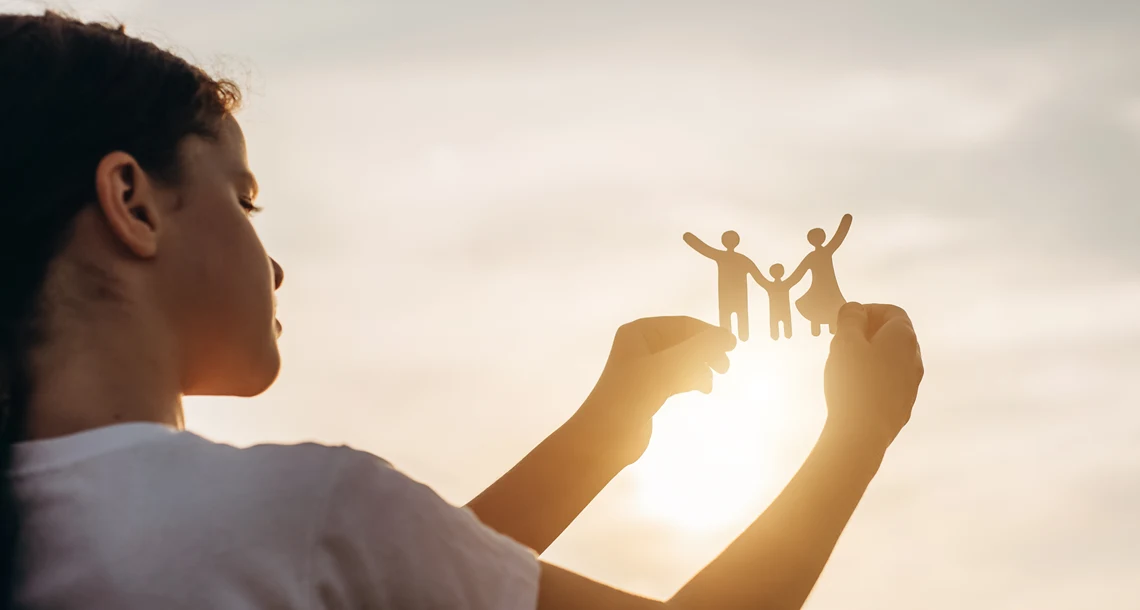 En flicka håller klippdockor i form av två vuxna med ett bar i mitten mot en himmel där solen bryter fram.