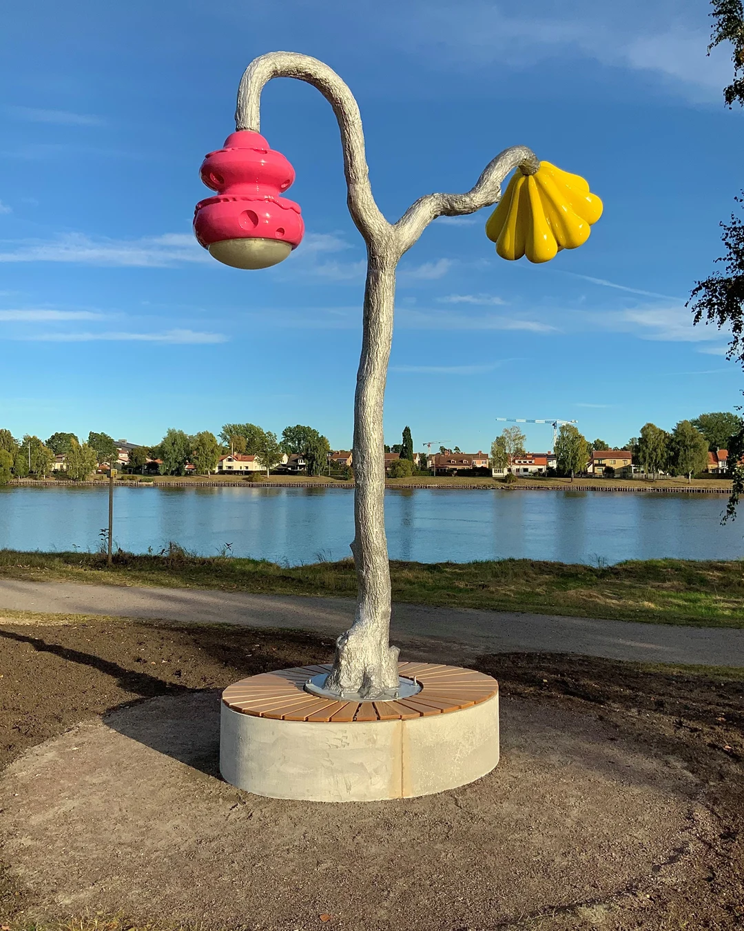 Ett konstverk utomhus besående av ett färgglatt träd av aluminium. Från den ena grenen växer en solgul blomma i plast, på den andra hänger en rosa bikupa i plast.