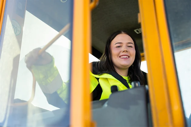 En glad tjej med gul reflexjacka sitter i en hjullastare.