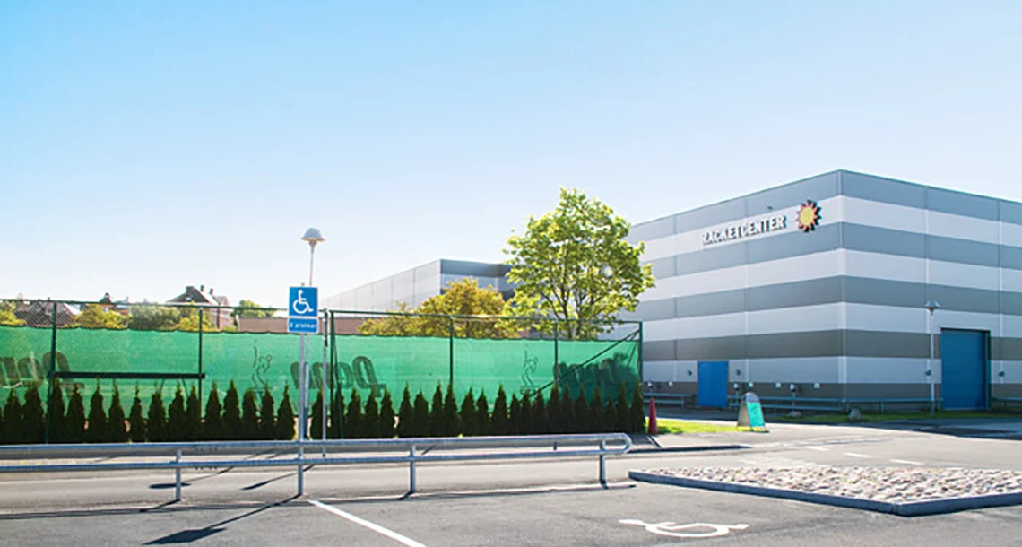 En handikapparkering framför ett grönt stängsel och en större byggnad med en sol och ett namn på väggen.