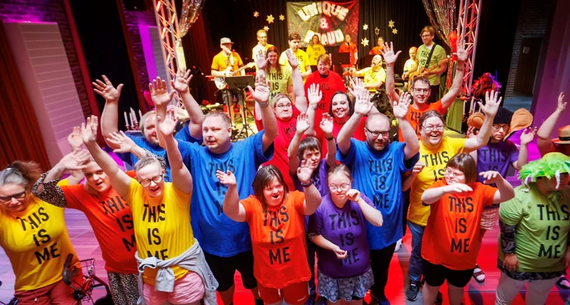 Personer i färgglada tröjor framför en scen. På tröjorna syns texten "This Is Me".
