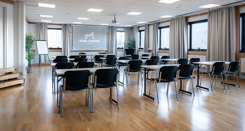 Flera mindre mötesbord med stolar står placerade i ett stort rum med brunt parkettgolv och är riktade mot en projektorskärm
