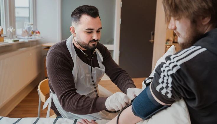En person övar på att ta blodtryck på en person som sitter på en säng i ett klassrum.