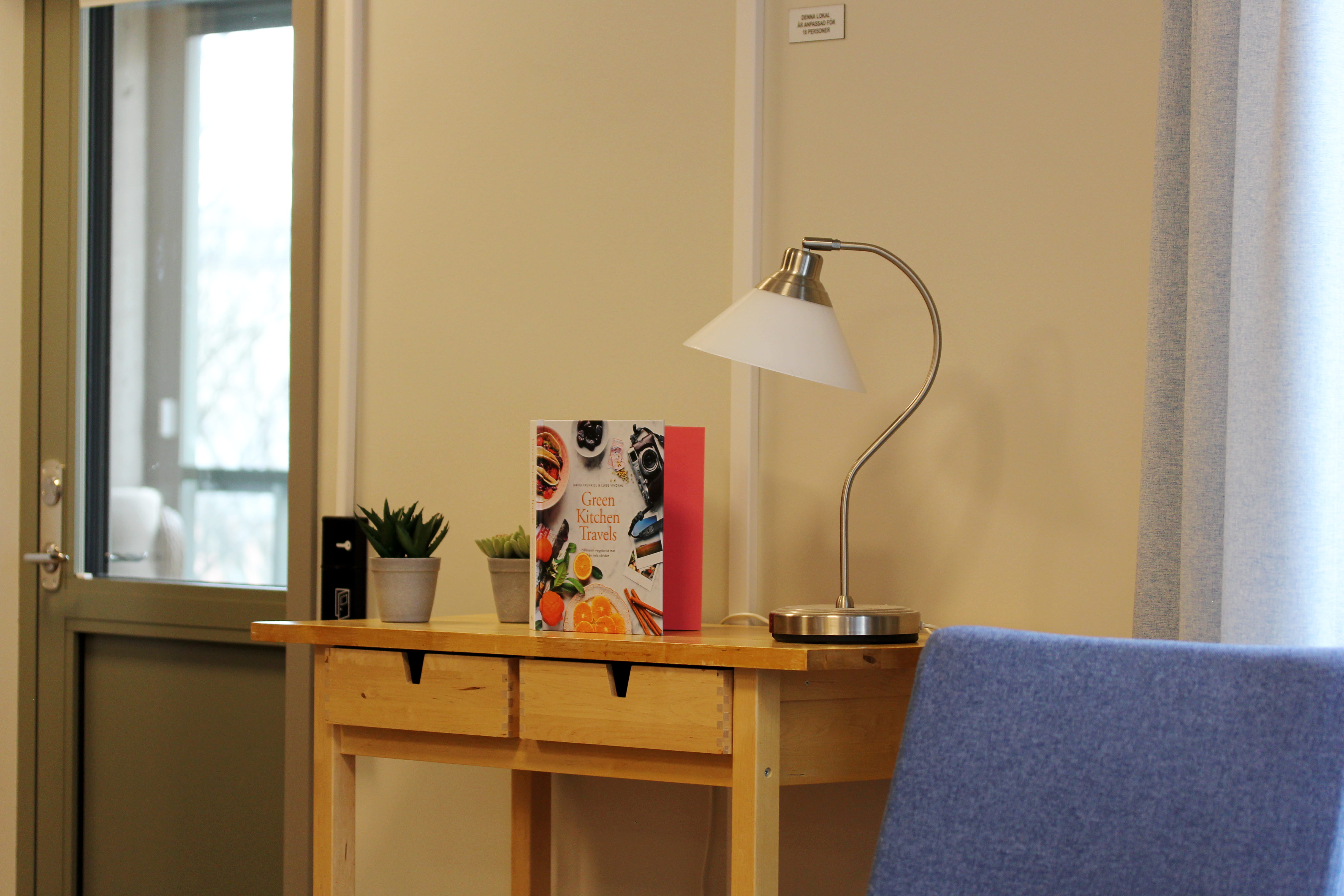 Ett avlastningsbord i trä med en bok, lampa och två små växter på i ett rum med vita väggar