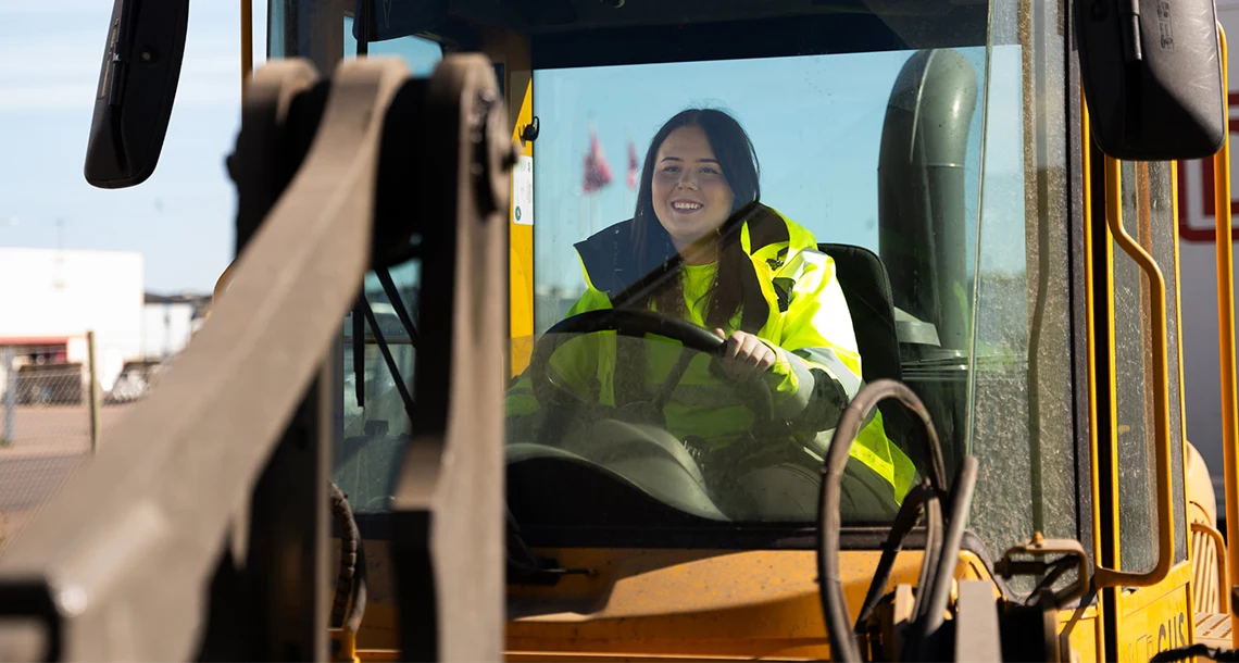 En glad tjej med gul reflexjacka sitter i och kör en hjullastare.