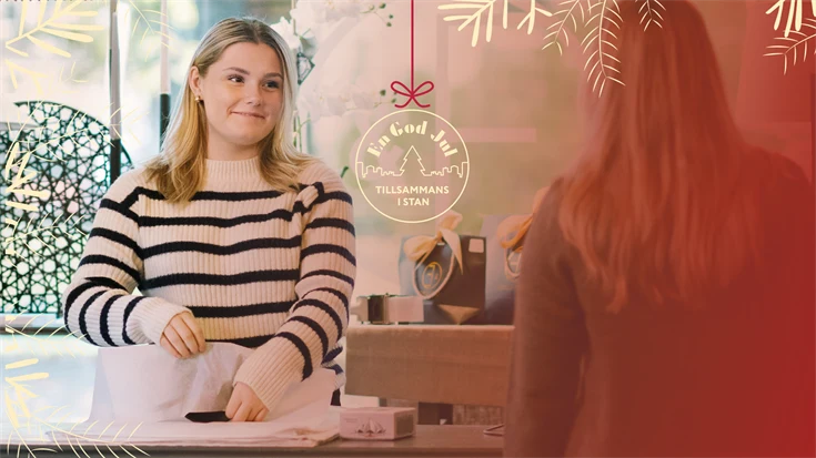 En person slår in en vara i vid en kassadisk i en inredningsbutik. En illustration av en julkula med texten "En god jul tillsammans i stan".