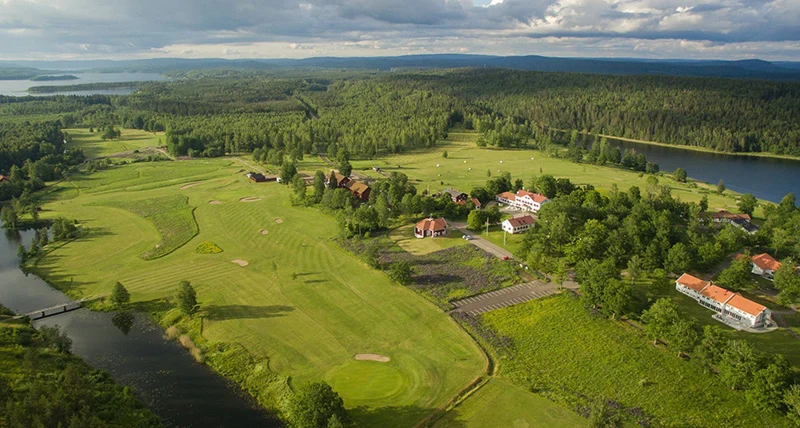 En flygbild över en grönskande golfbana med hus, skog och vatten som omgärdar banan.
