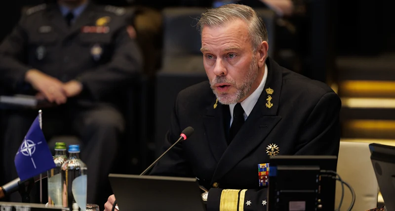 En person i en mörk militär uniform sitter vid ett konferensbord med mikrofon framför sig. På bordet finns en NATO-flagga, vattenflaskor, en laptop och teknisk utrustning. I bakgrunden syns andra uniformerade personer sitta och lyssna i en större möt