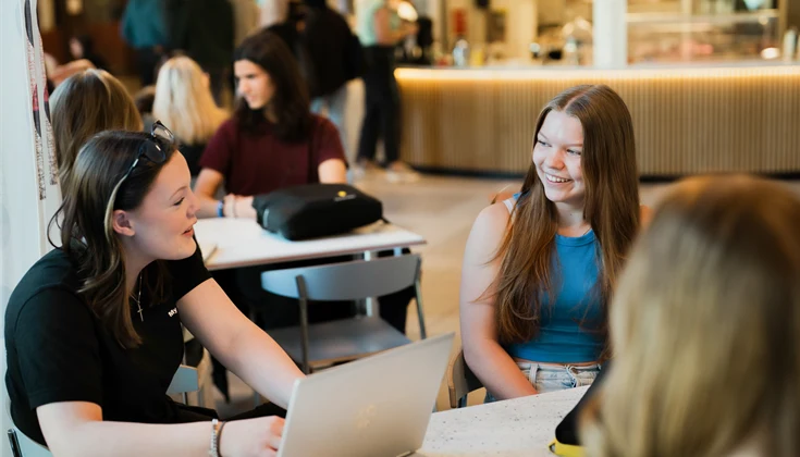 Tre elever sitter och pluggar tillsammans i en kafeteria. Fler elever syns i bakgrunden.