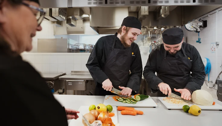 Tre personer hackar grönsaker i ett restaurangkök.
