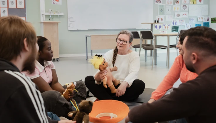 Fem personer sitter på golvet i ett klassrum i en ring och håller i gosedjur.