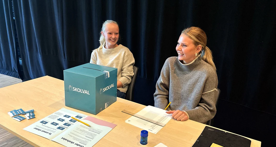 Två gymnasieelever sitter vid ett bord där röster lämnas in i riksdagsskolvalet.