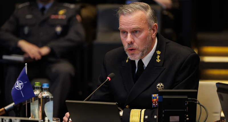 En person i en mörk militär uniform sitter vid ett konferensbord med mikrofon framför sig. På bordet finns en NATO-flagga, vattenflaskor, en laptop och teknisk utrustning. I bakgrunden syns andra uniformerade personer sitta och lyssna i en större möt
