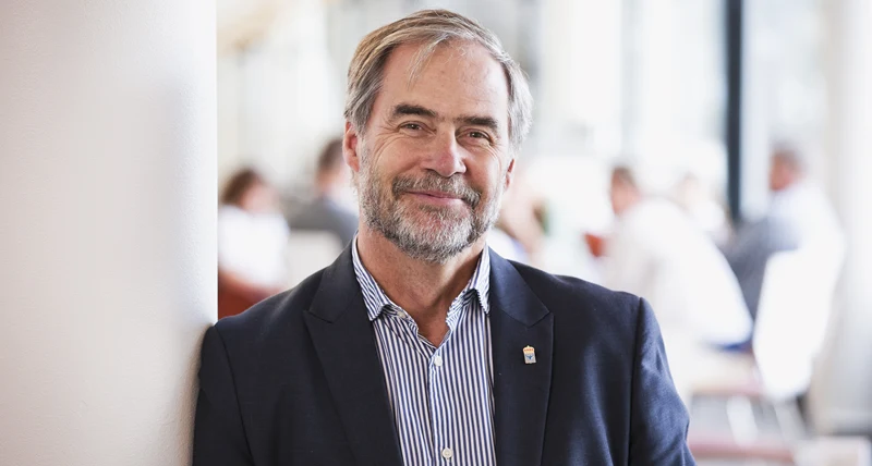 Man i kavaj och randig skjorta lutar sig mot en stolpe inomhus och ler mot kameran.