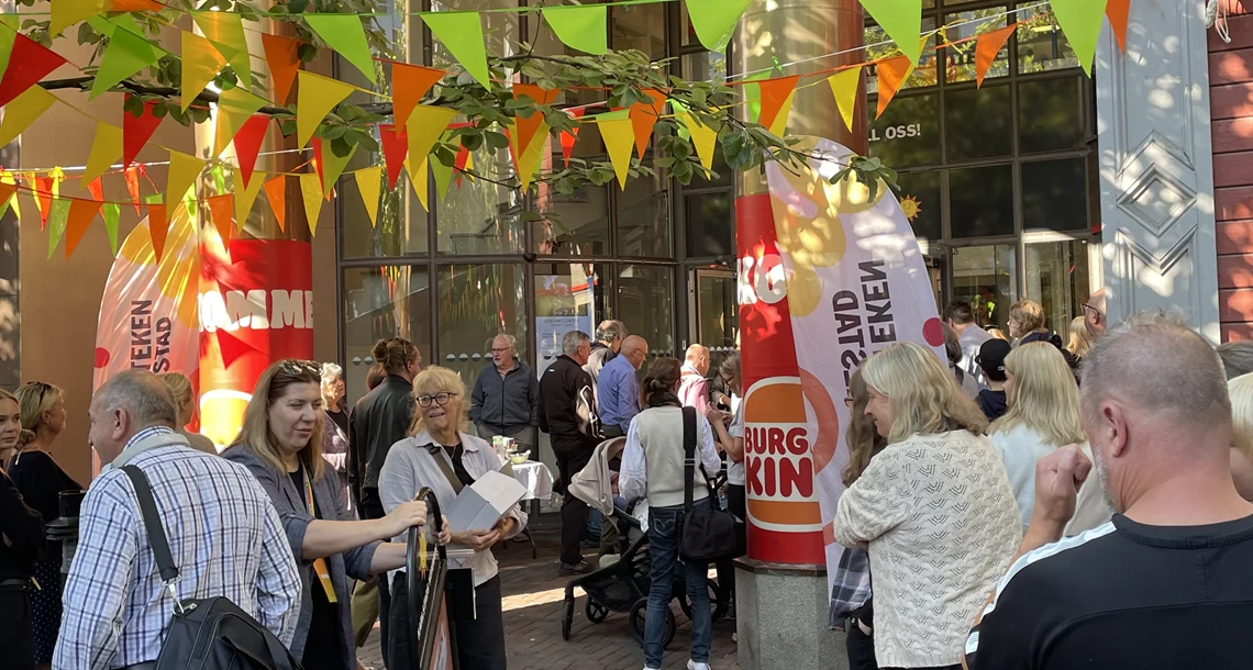 Folksamling utanför byggnad. Vimplar och träd i förgrunden.
