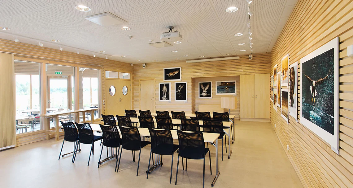 Flera mindre mötesbord med stolar står placerade i ett rum med ljusbruna färger och naturmaterial.rskärm