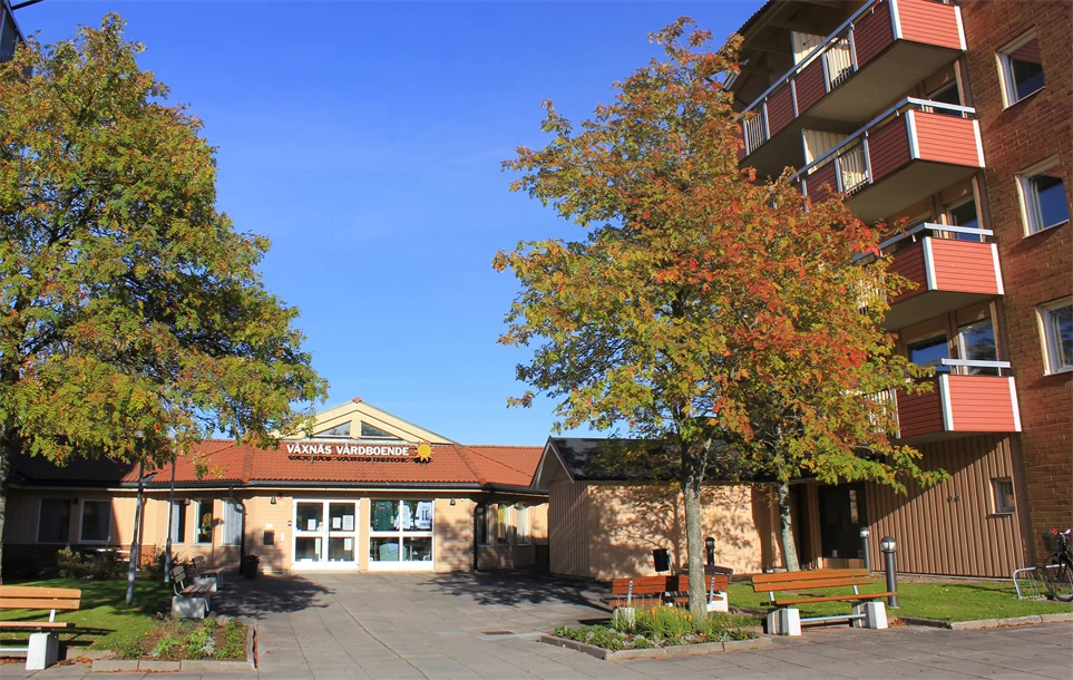 Våxnäs trygghetsboende med en molnfri och blå himmel bakom. På varsin sida om entrén står två stora träd i höstfärgerna orange, rött och grönt. Ovanför dörren står texten "Våxnäs vårdboende" med karlstadsolen på slutet.