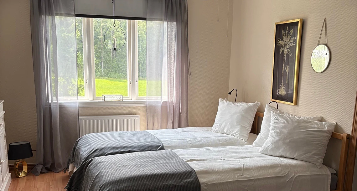 Två bäddar i ett rum med utsikt genom ett fönster mot ett skogsbryn
