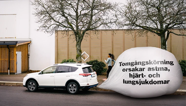 En bil står framför en byggnad och går på tomgång. Bakom bilen ser man en bubbla av avgaser. I den bubblan står texten "Tomgångskörning orsakar astma, hjärt- och kärlsjukdomar ".