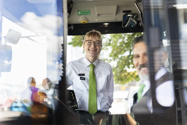 Två bussförare tittar ut genom ett fönster i en buss och ler.