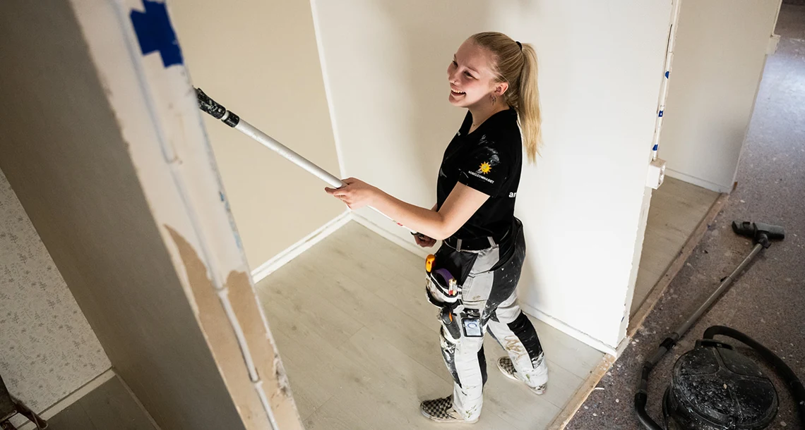 Glad tjej använder en roller på ett skaft för att måla vit färg på en vägg.