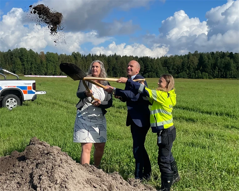 Två kvinnor och en man håller i en spade med tre handtag och tar ett gemensamt spadtag.