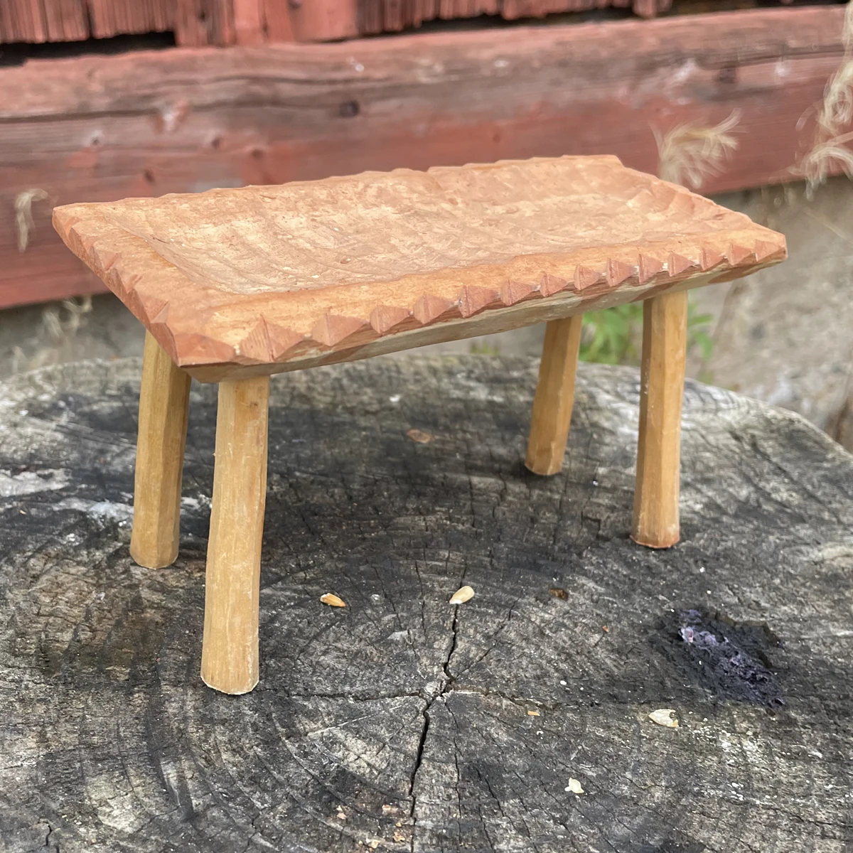 Knivslöjdad pall i trä på stubbe framför faluröd vägg