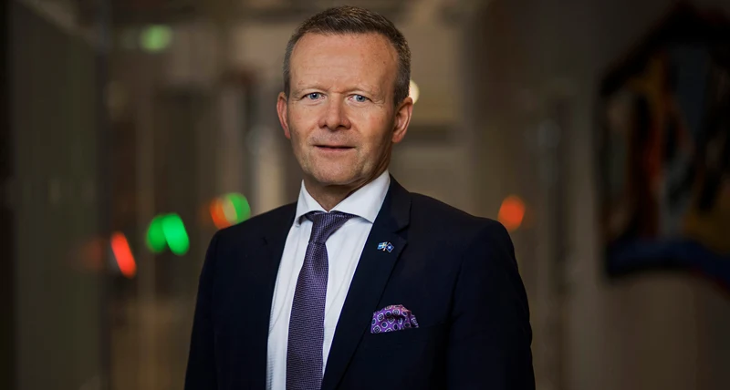 Man med svart kostym och lila slips står inomhus och ler mot kameran