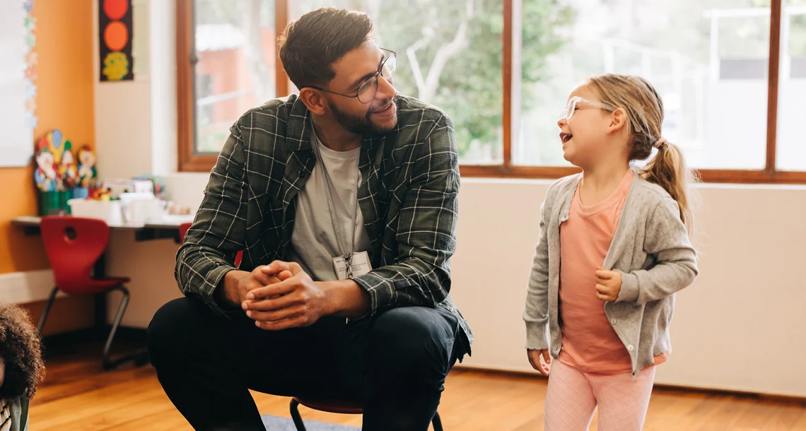 En barnskötare sitter på en stol och ler åt ett barn i ett klassrum.