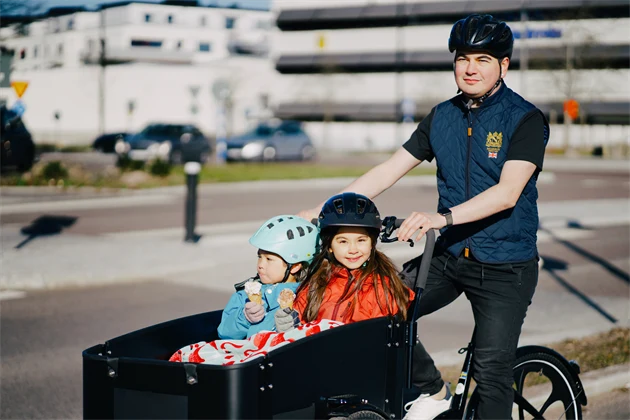Två barn i en lastcykel som äter glass. En pappa som kör dem. 
