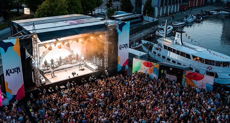 En utomhusscen där en konsert pågår md en stor mängd publik framför och en hamn med båtar i bakgrunden.