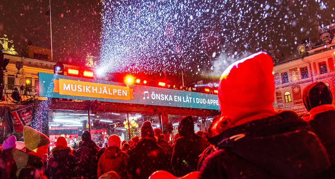 En samling människor som står framför en glasbur med texten musikhjälpen på