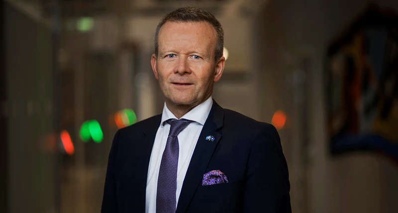Man med svart kostym och lila slips står inomhus och ler mot kameran