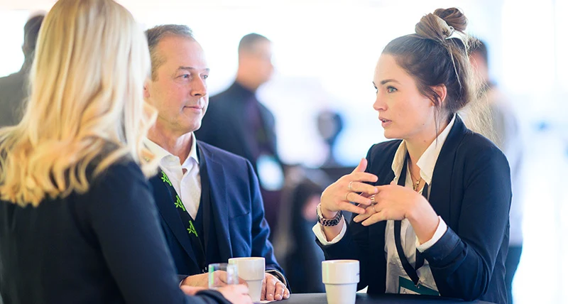 Två kvinnor och en man står vid ett bord med kaffekoppar och samtalar.