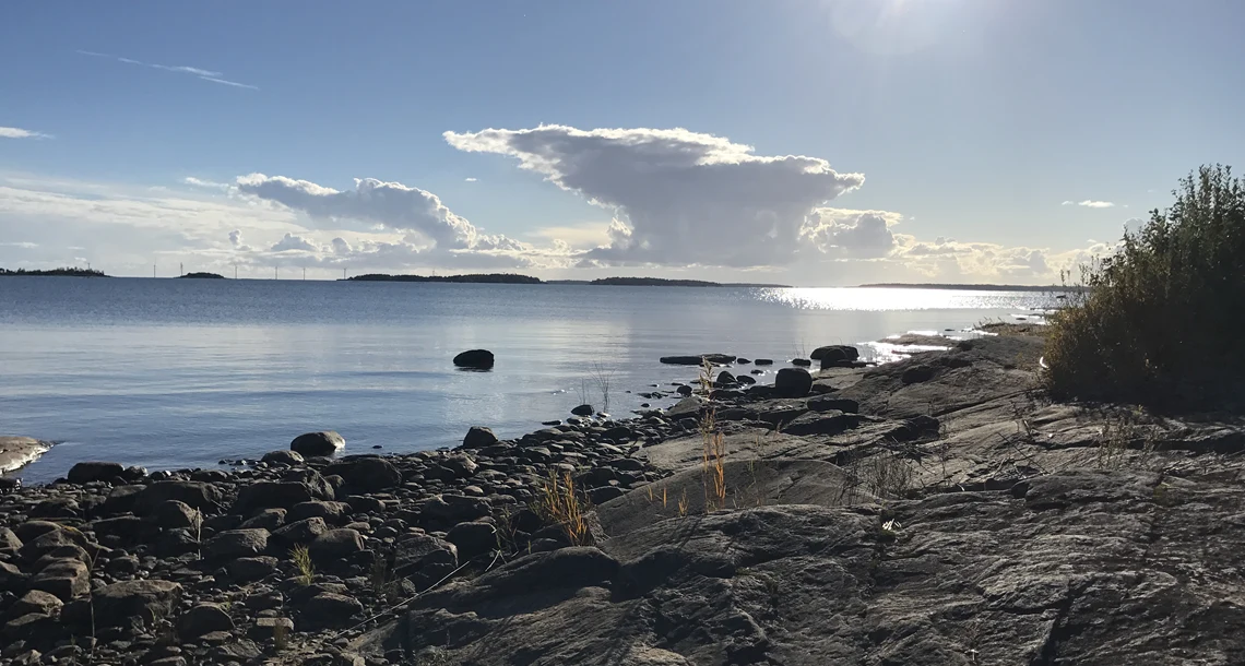 Solig kustvy med klippor i förgrunden, stilla vatten som sträcker sig mot horisonten och små öar 